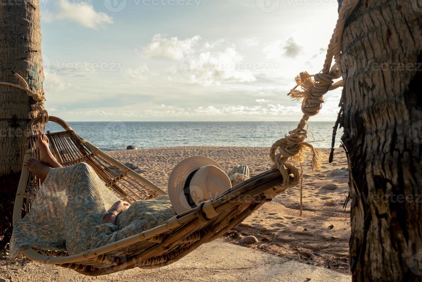 skön ung asiatisk kvinna med hatt avkopplande avkopplande i en hängmatta mellan handflatan träd, sommar Lycklig strand semester begrepp. foto