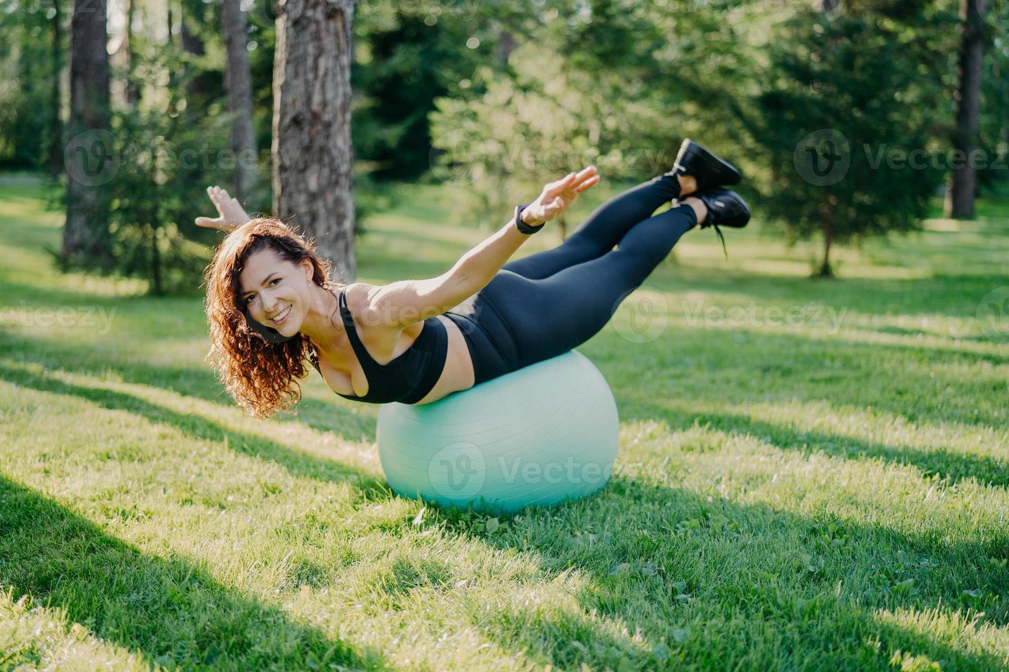 leende glad brunett kvinna med europeiskt utseende balanserar på träningsboll sträcker ut armarna klädda i topp och leggings poserar utomhus i sommarparken och gör pilates i naturen under solig dag foto