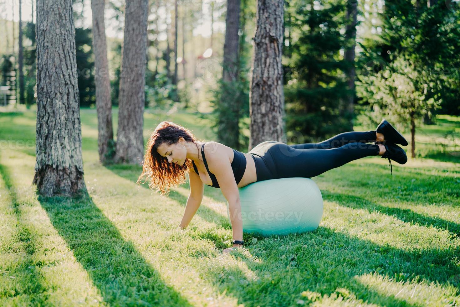 utomhus skott av sportig ung kvinna lutar sig mot fitness bollen håller balansen och gör gymnastiska övningar bär avskurna leggings sneakers har träning i skogen poser på grönt gräs nära träd. foto