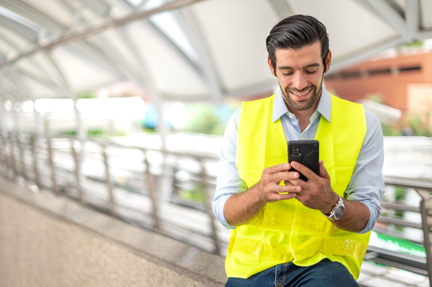 stänga upp caucasian man använda sig av en smartphone för kommunicera med hans medarbetare och för Kontakt med hans vän medan han bär gul väst och arbetssätt på hans webbplats. foto