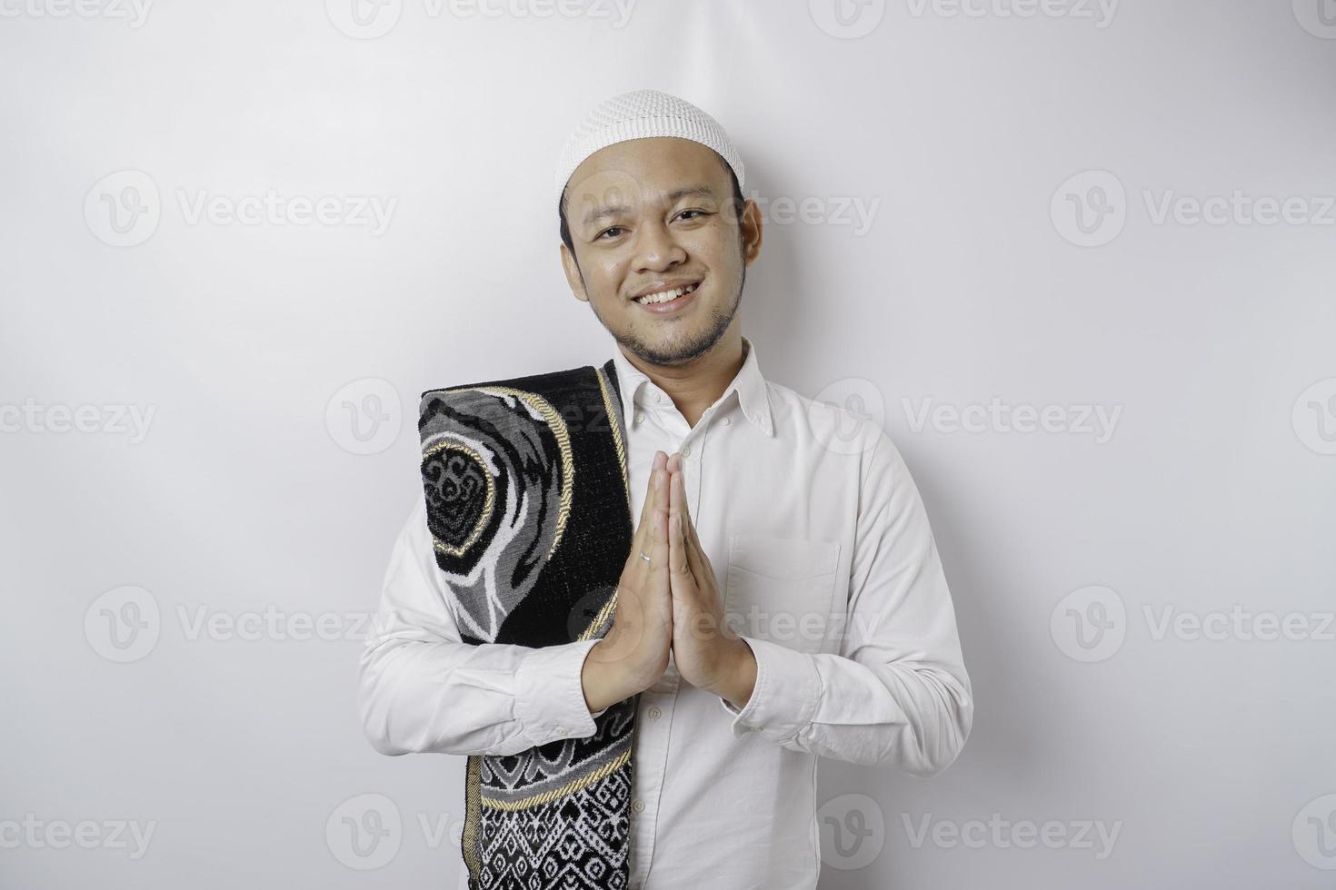 leende ung asiatisk muslim man med bön matta på hans axel, gestikulerar traditionell hälsning isolerat över vit bakgrund foto