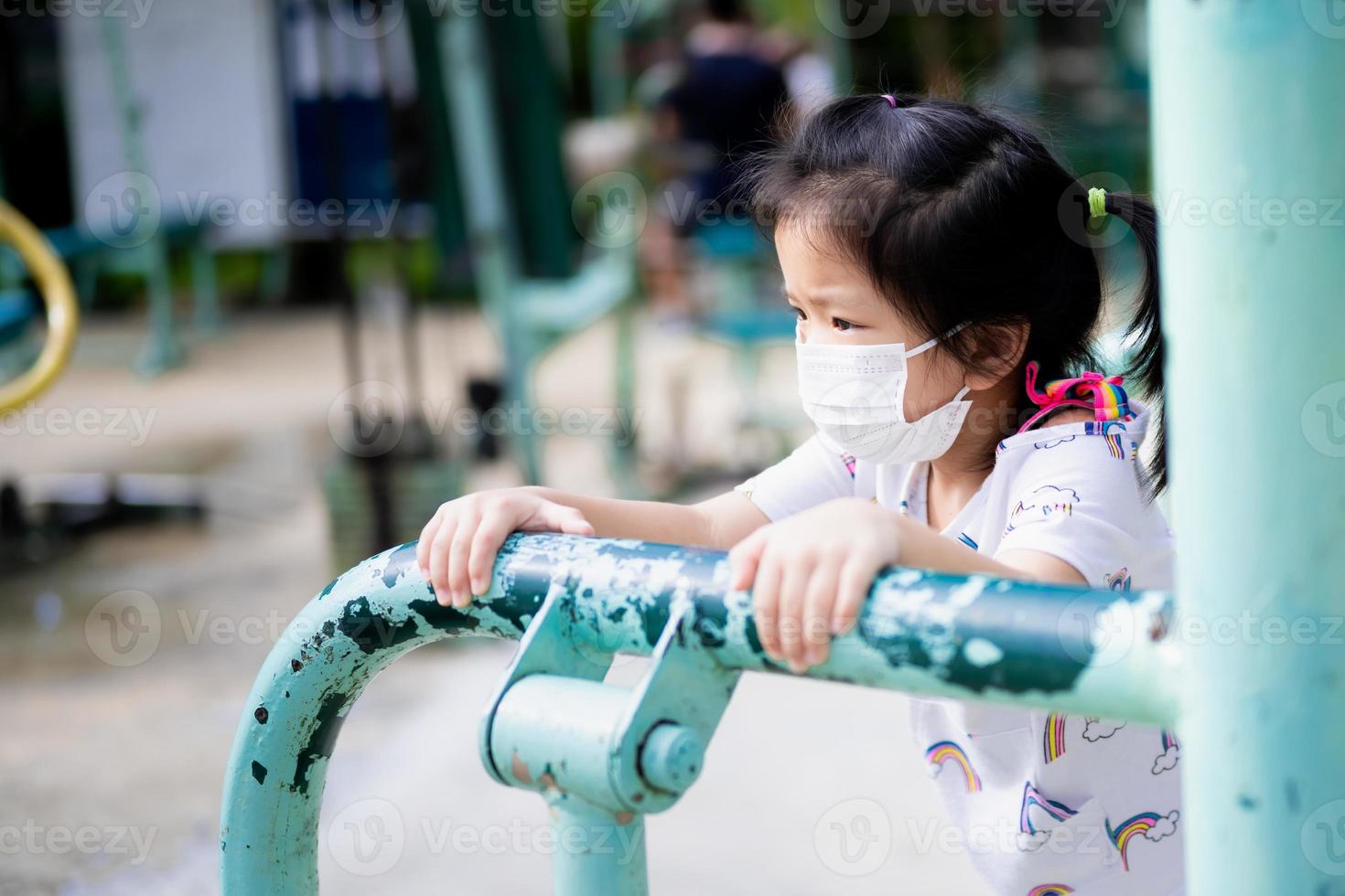 porträtt söt unge flicka spelar övning Utrustning på lekplats. barn bär vit medicinsk ansikte mask. förhindra toxisk damm PM 2,5 och spridning coronavirus covid19. tömma Plats. foto
