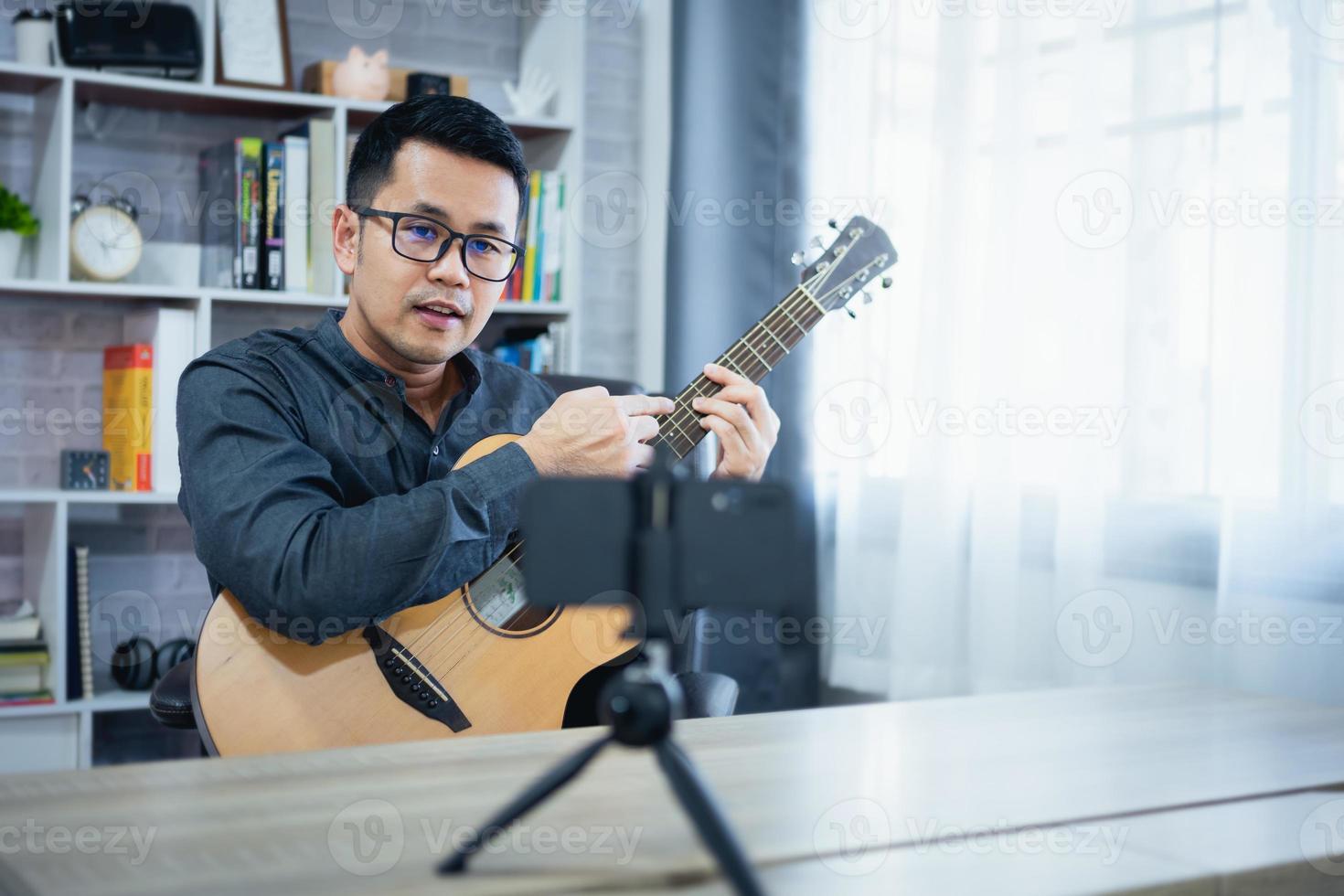 asiatisk musik lärare man sändningar leva gitarr lektioner till studenter uppkopplad använder sig av mobil. asiatisk manlig musiker spelar gitarr sång leva video konferenser på Hem. leva konsert musik från var som helst. foto