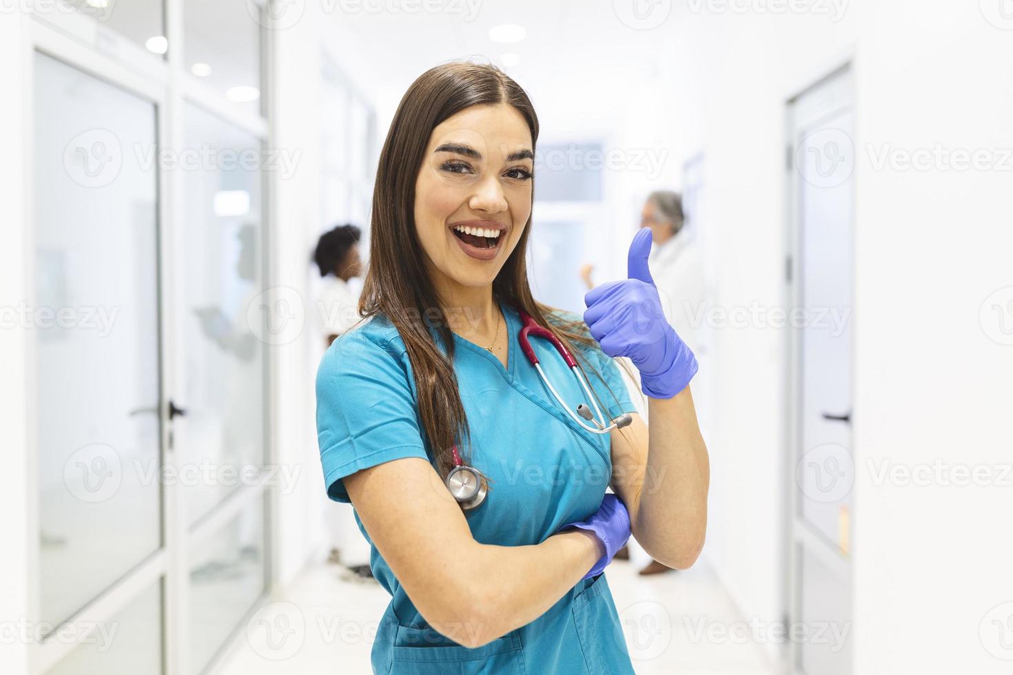 porträtt av leende kvinna läkare bär scrubs i upptagen sjukhus korridor som visar tummen upp foto