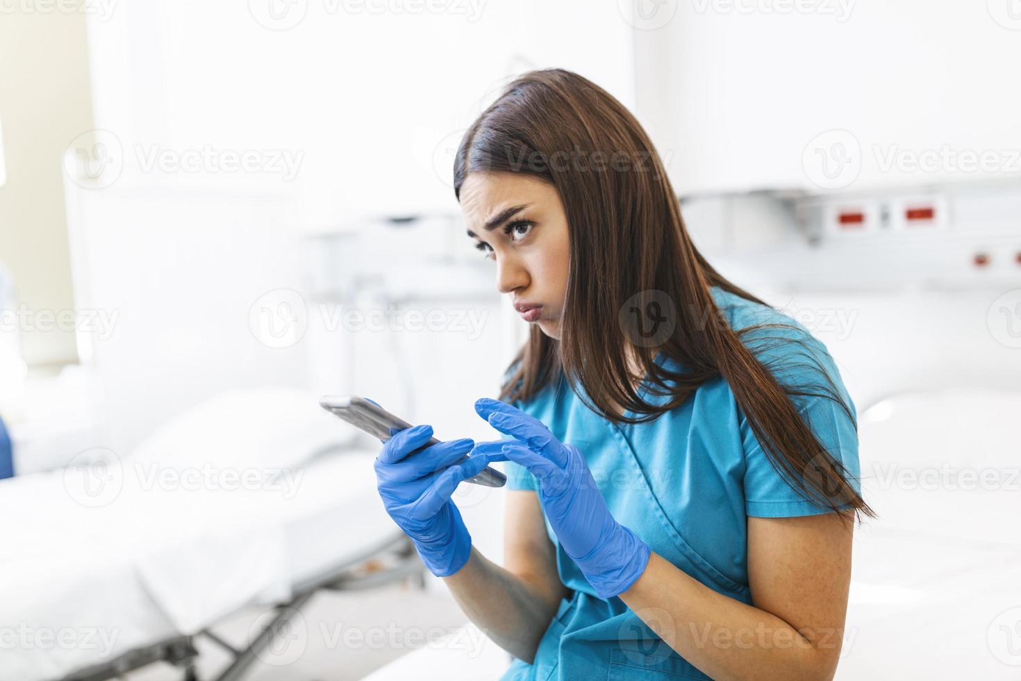 ett utmattad kvinna sjuksköterska sitter på en säng i en sjukhus under en flytta ha sönder. hon dagdrömmar medan innehav hans smartphone. foto