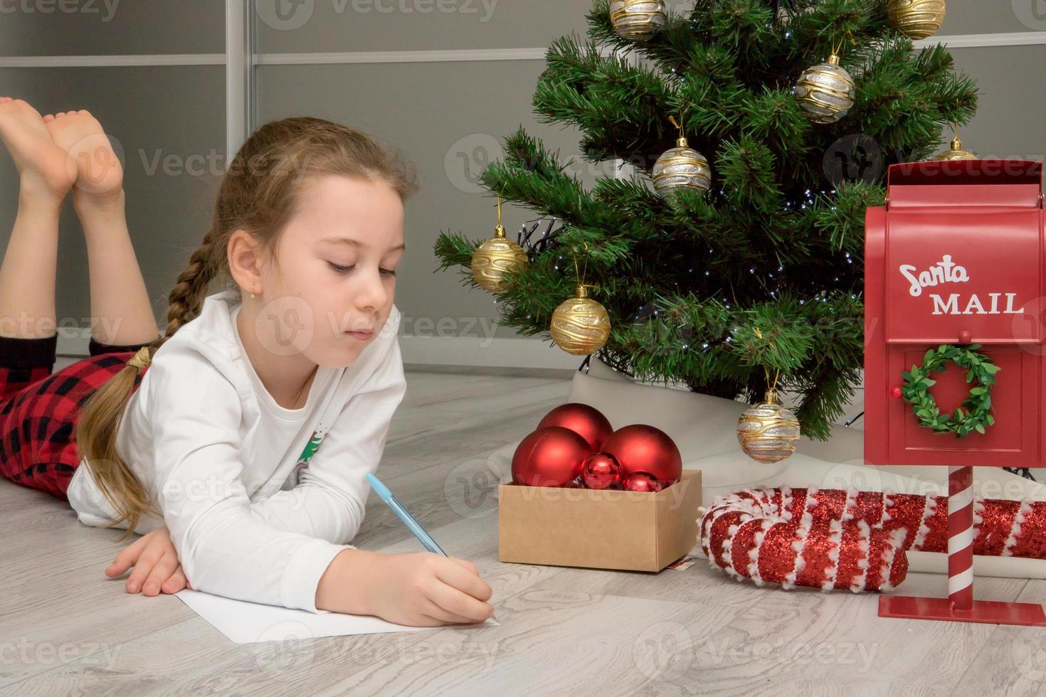 flicka i pyjamas på Hem nära de jul träd med koncentration skrivningar en brev till tomte, jul innehåll foto