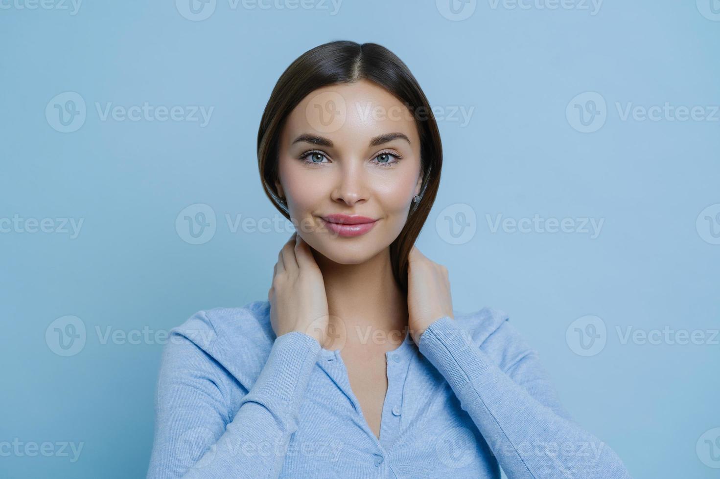 headshot av ung härlig kvinna håller händerna på halsen, har europeiskt utseende, glad att höra trevliga ord, bär casual tröja, isolerad över blå bakgrund. mänskligt ansikte uttryck koncept foto