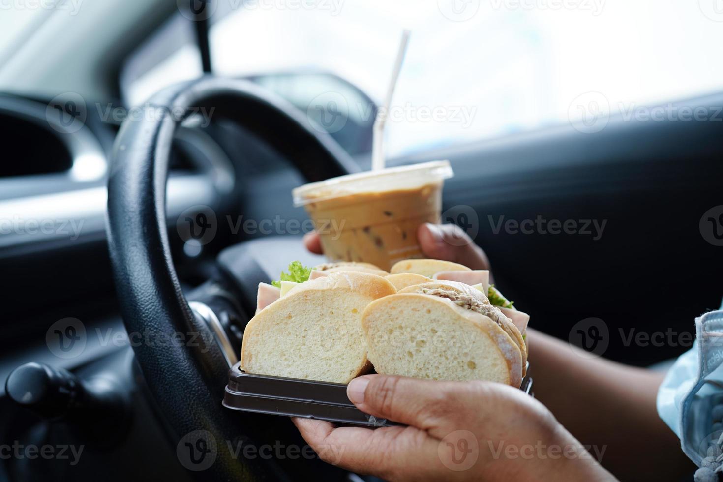 asiatisk kvinna förare håll is kaffe kopp och smörgås bröd för äta och dryck i bil, farlig och risk ett olycka. foto