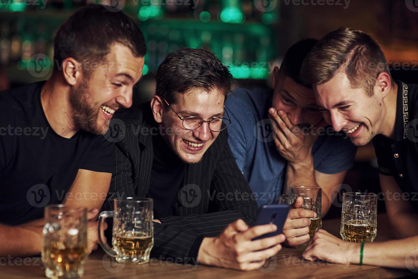 rolig innehåll på smartphone. tre vänner vilar i de pub med öl i händer. har konversation foto