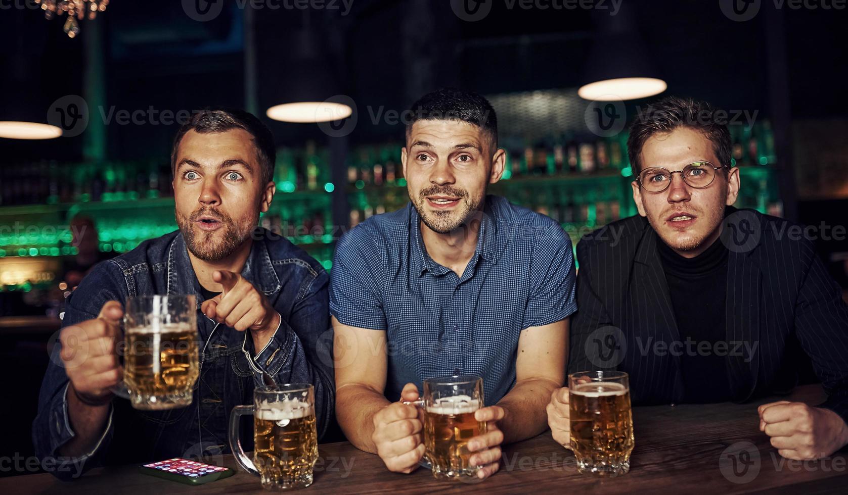 tre sporter fläktar i en bar tittar på fotboll. med öl i händer foto