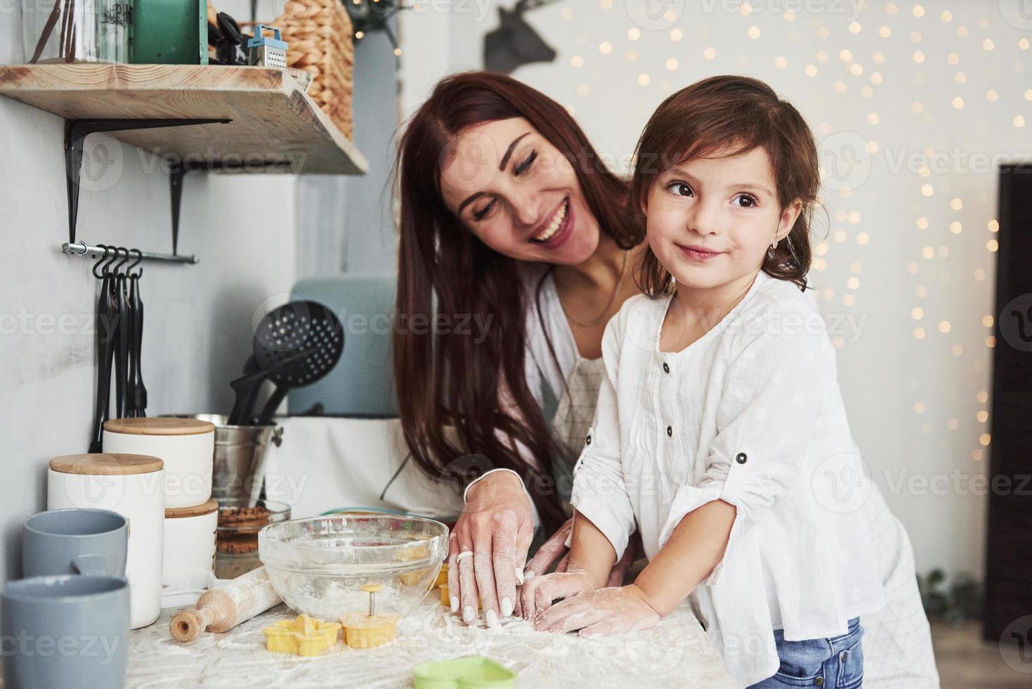 ser till de sida medan kvinna är leende. Lycklig dotter och mamma är framställning bageri Produkter tillsammans. liten hjälpare i de kök foto