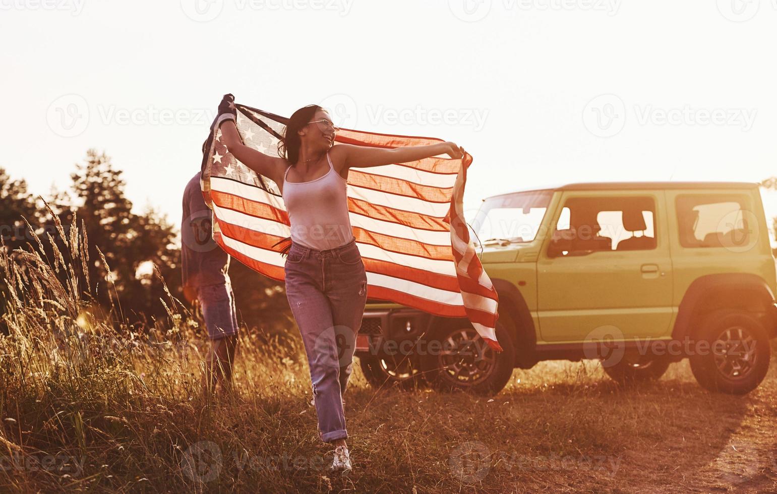 flicka kör fram. vänner ha trevlig helgen utomhus nära deras grön bil med USA flagga foto