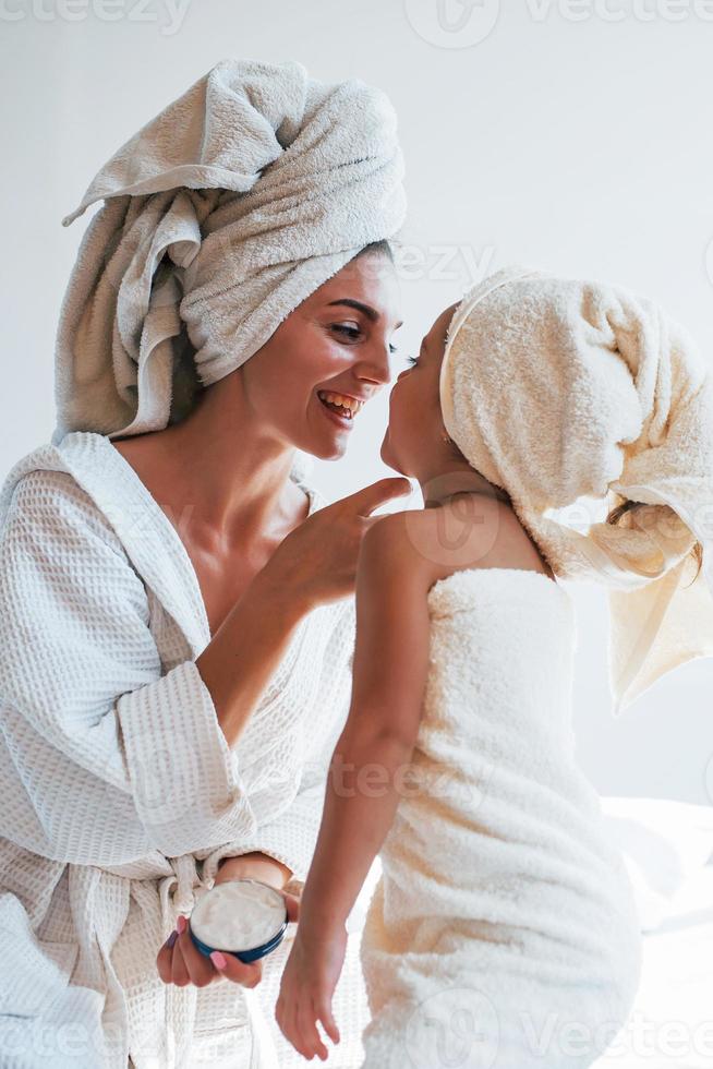 använder sig av grädde till klar hud. ung mor med henne dotter ha skönhet dag inomhus i vit rum foto