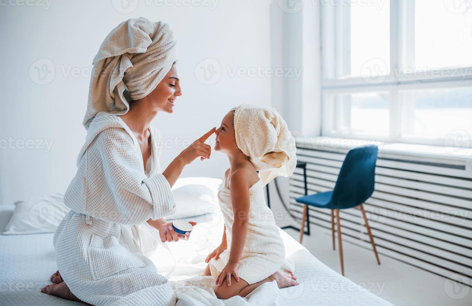 använder sig av grädde till klar hud. ung mor med henne dotter ha skönhet dag inomhus i vit rum foto