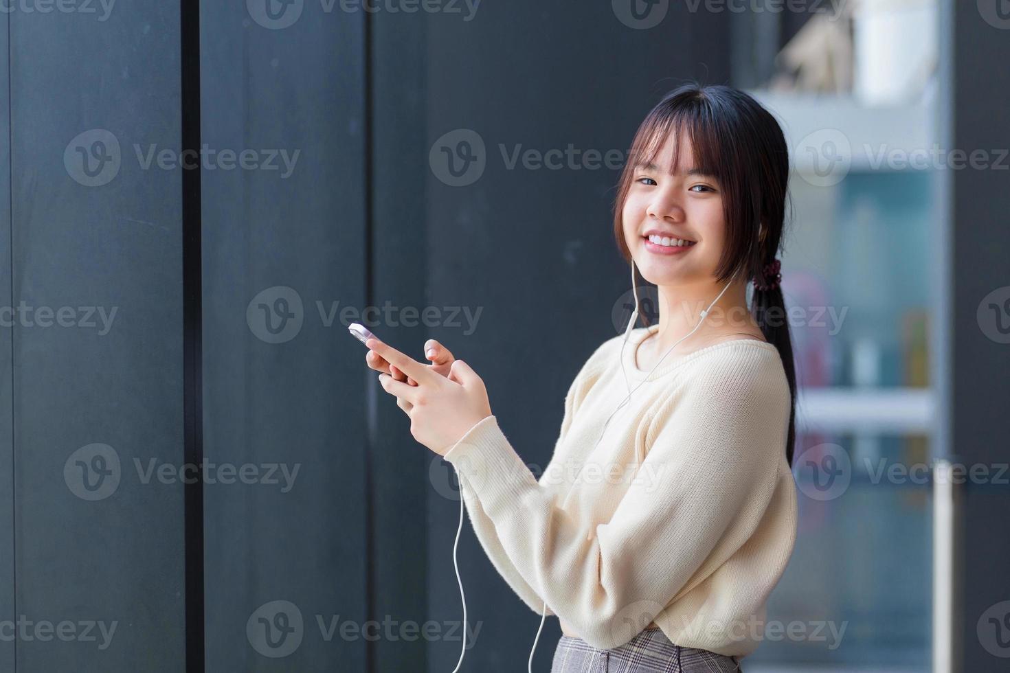 söt ung asiatisk studerande bär en långärmad skjorta är stående och leende medan använder sig av henne smartphone och hörlurar till lyssna en föreläsning självsäkert som hon väntar till stiga på de klassrum på skola. foto