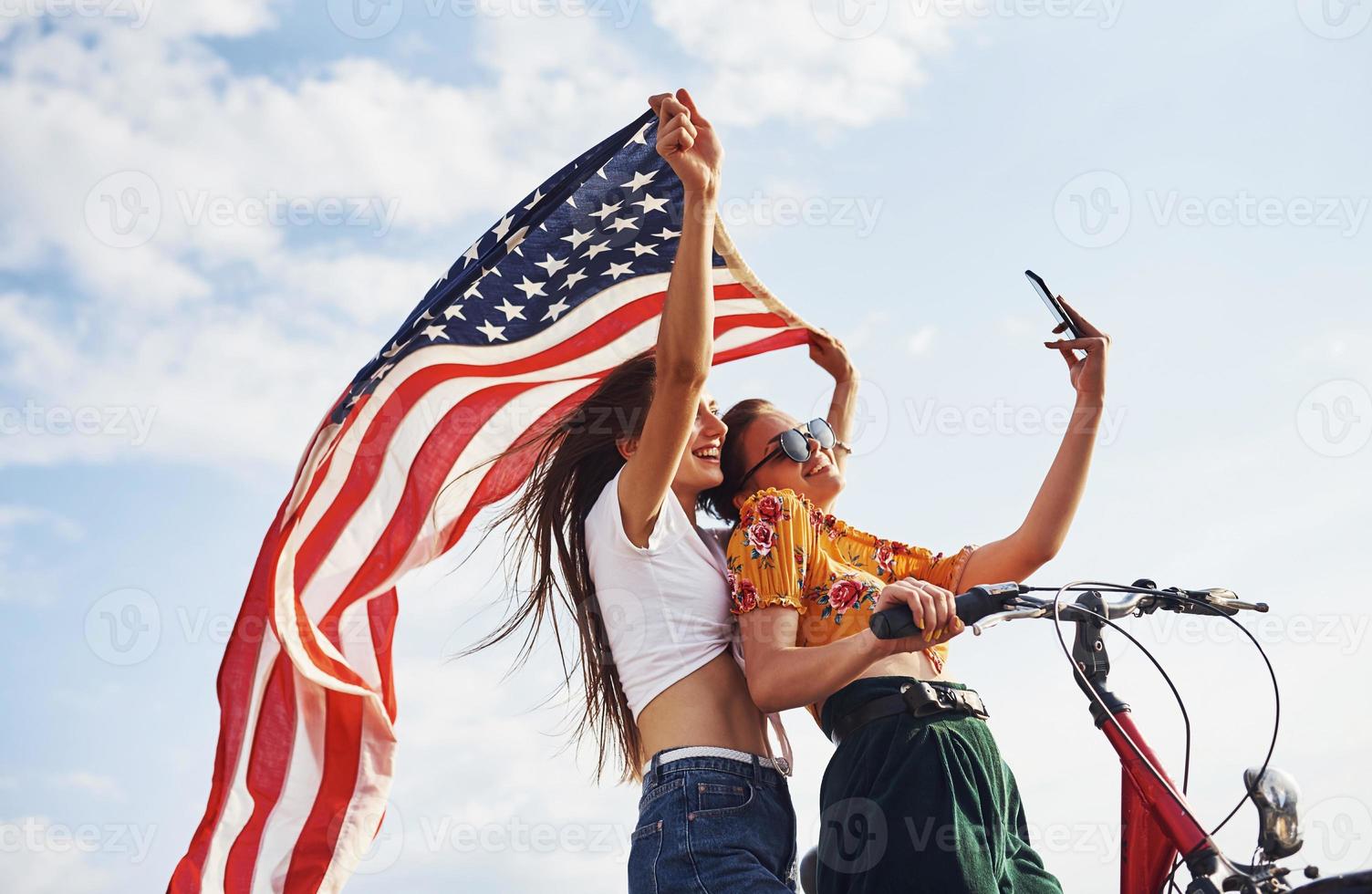 två patriotisk glad kvinnor med cykel och USA flagga i händer gör selfie foto