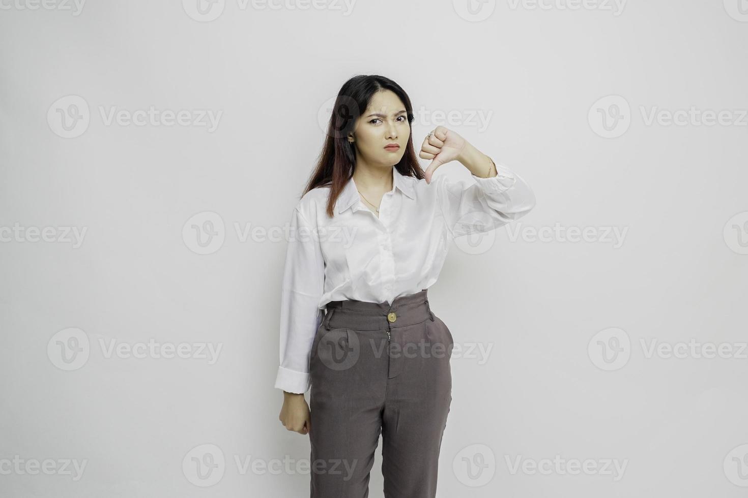 besviken asiatisk kvinna bär vit skjorta ger tummen ner hand gest av godkännande, isolerat förbi vit bakgrund foto