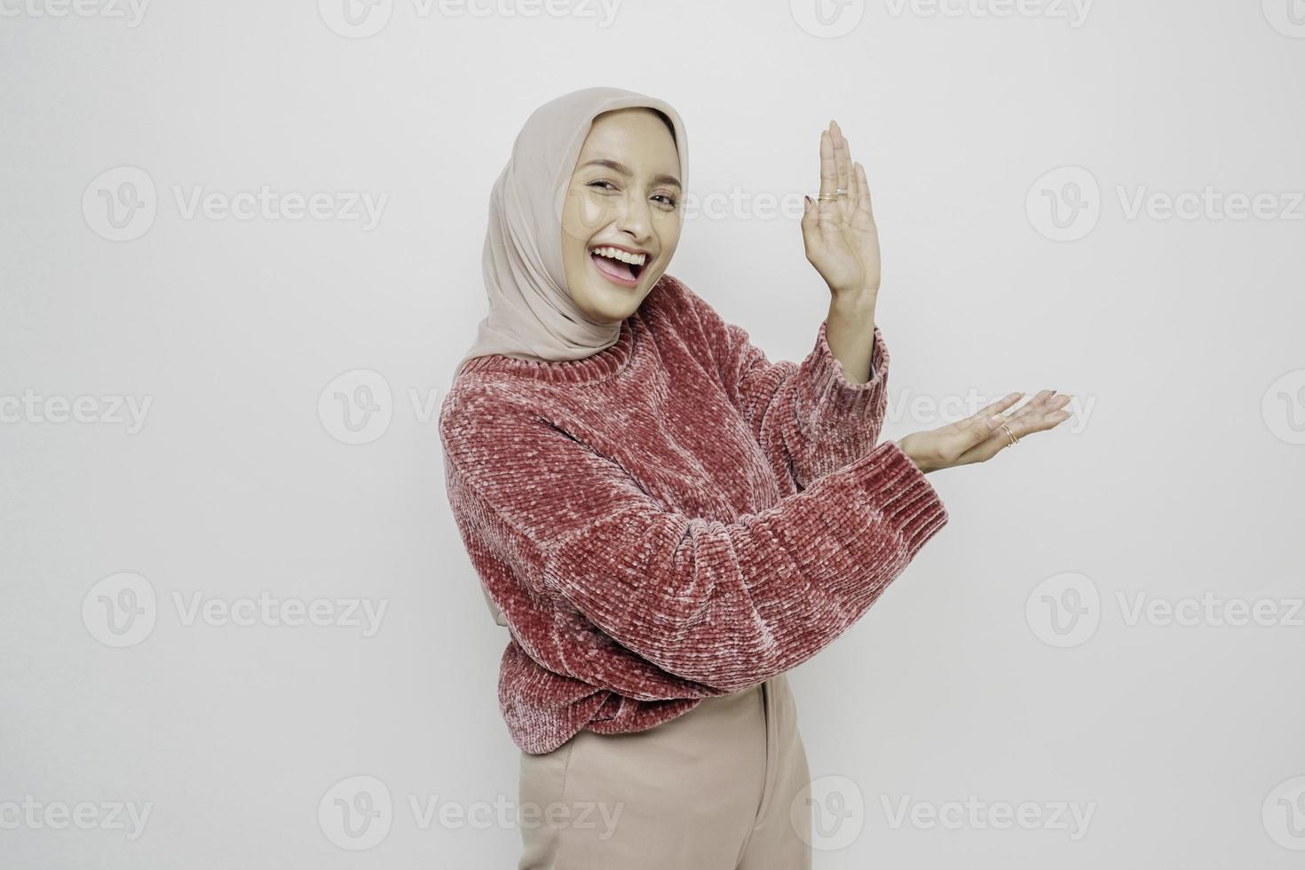 upphetsad asiatisk muslim kvinna bär rosa Tröja och hijab pekande på de kopia Plats bredvid henne, isolerat förbi vit bakgrund foto