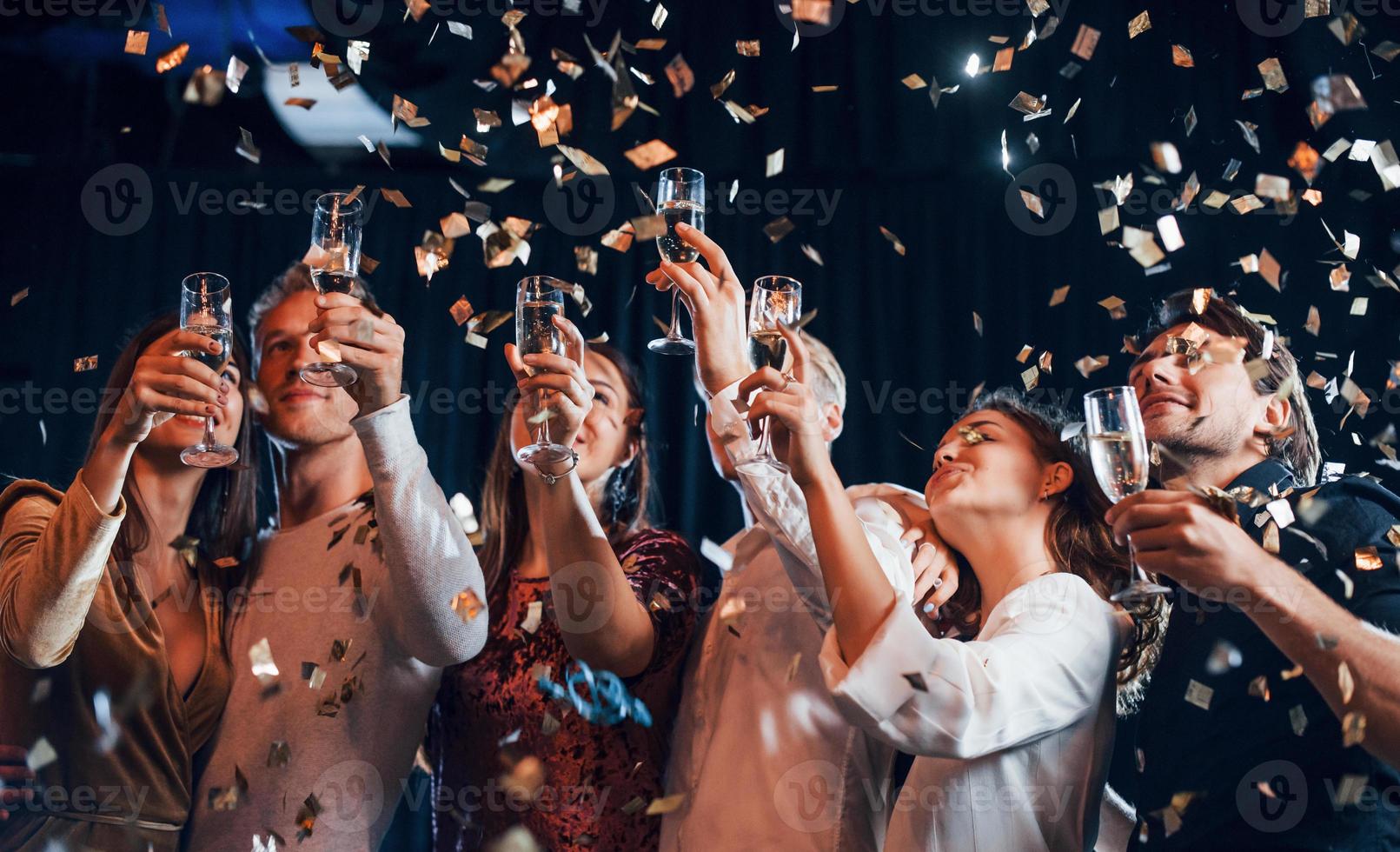 konfetti är i de luft. grupp av glad vänner fira ny år inomhus med drycker i händer foto