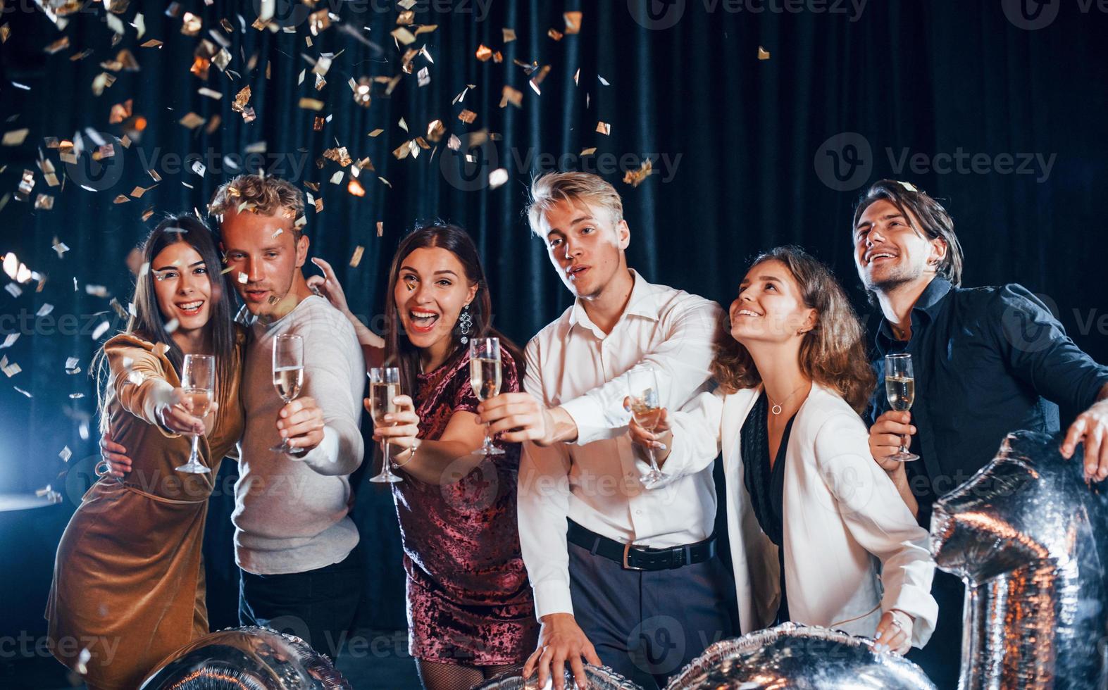 konfetti är i de luft. grupp av glad vänner fira ny år inomhus med drycker i händer foto