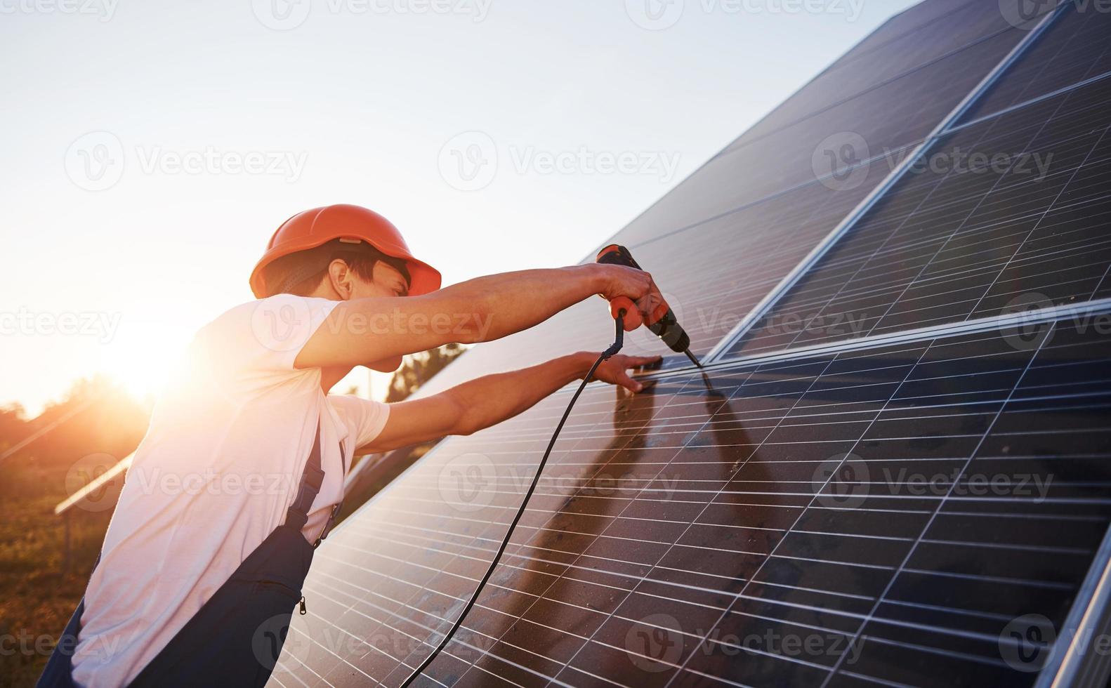 använder sig av sladdlös skruvmejsel. manlig arbetstagare i blå enhetlig utomhus med sol- batterier på solig dag foto