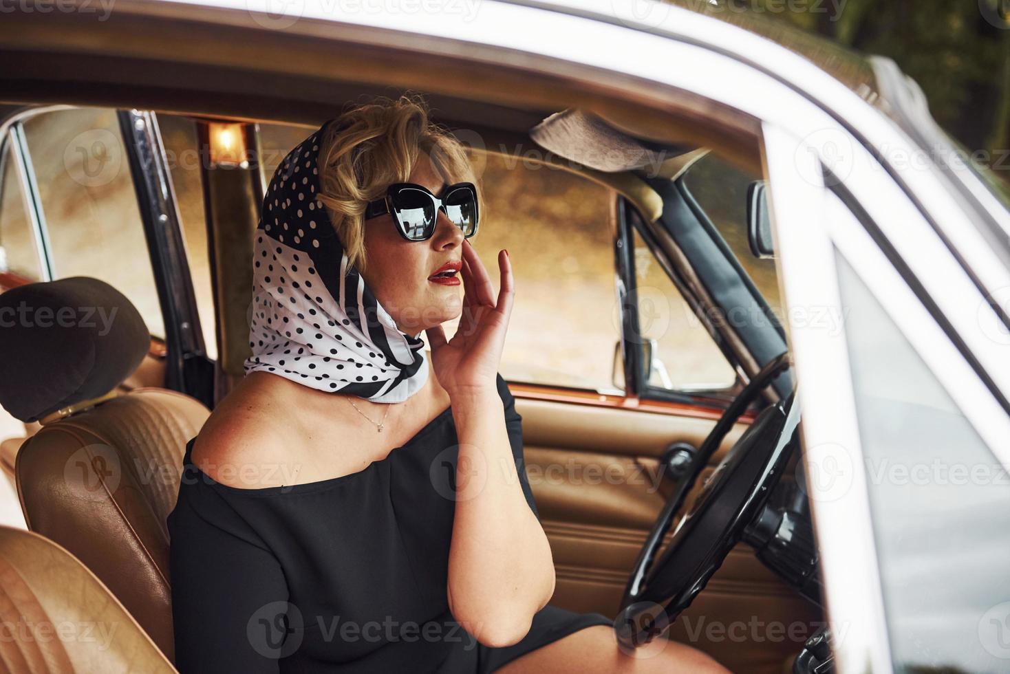 blond kvinna i solglasögon och i svart klänning sitter i gammal årgång klassisk bil foto