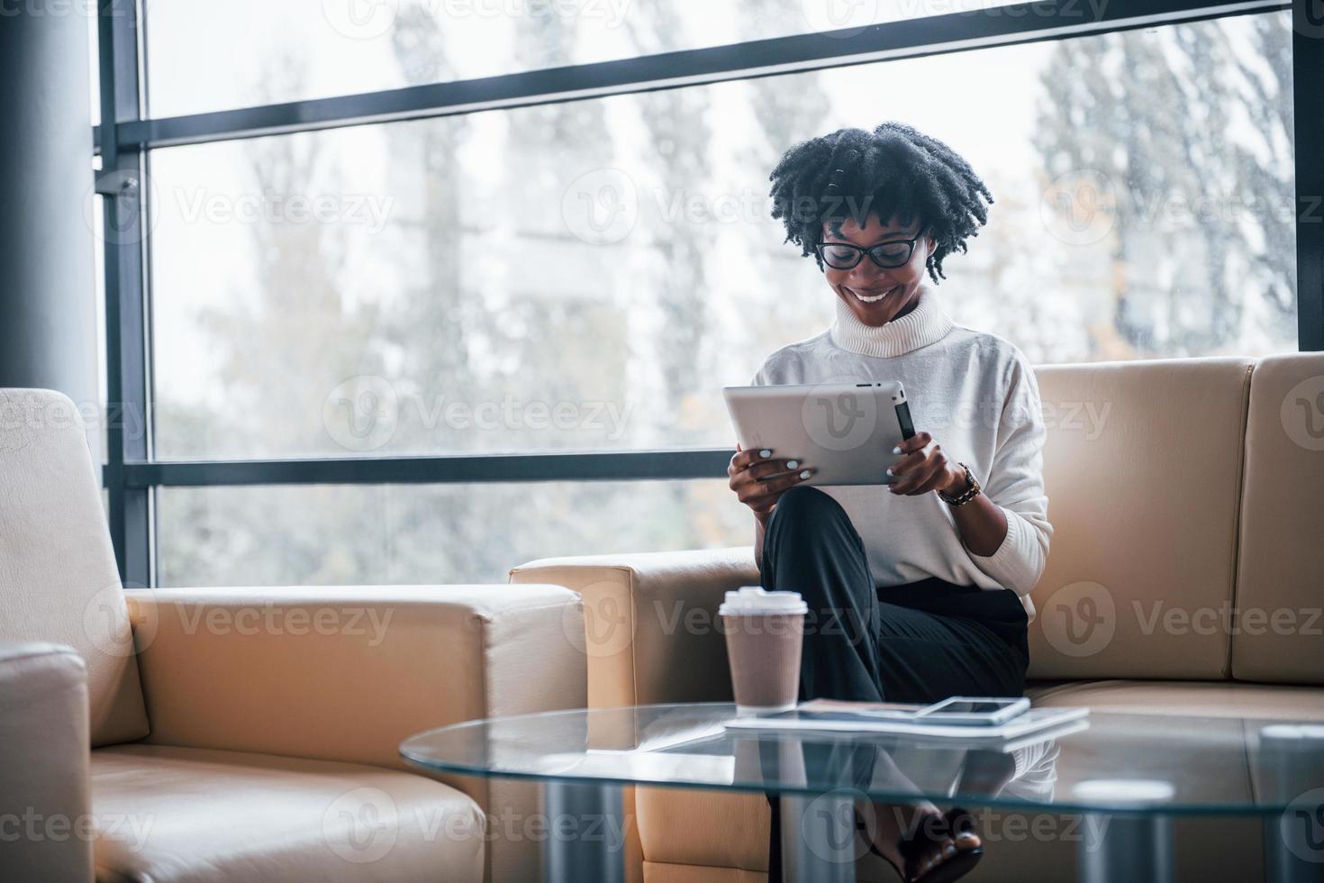 ung afrikansk amerikan kvinna i glasögon sitter inomhus i de kontor med läsplatta i händer foto