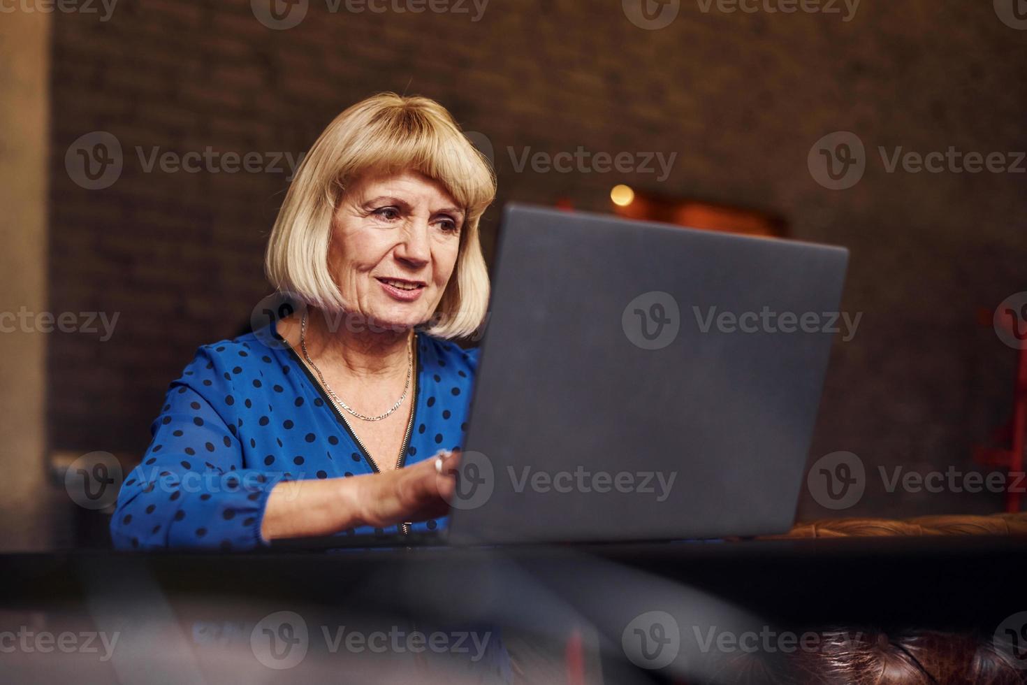 porträtt av senior kvinna den där sitter inomhus i de Kafé med modern bärbar dator foto