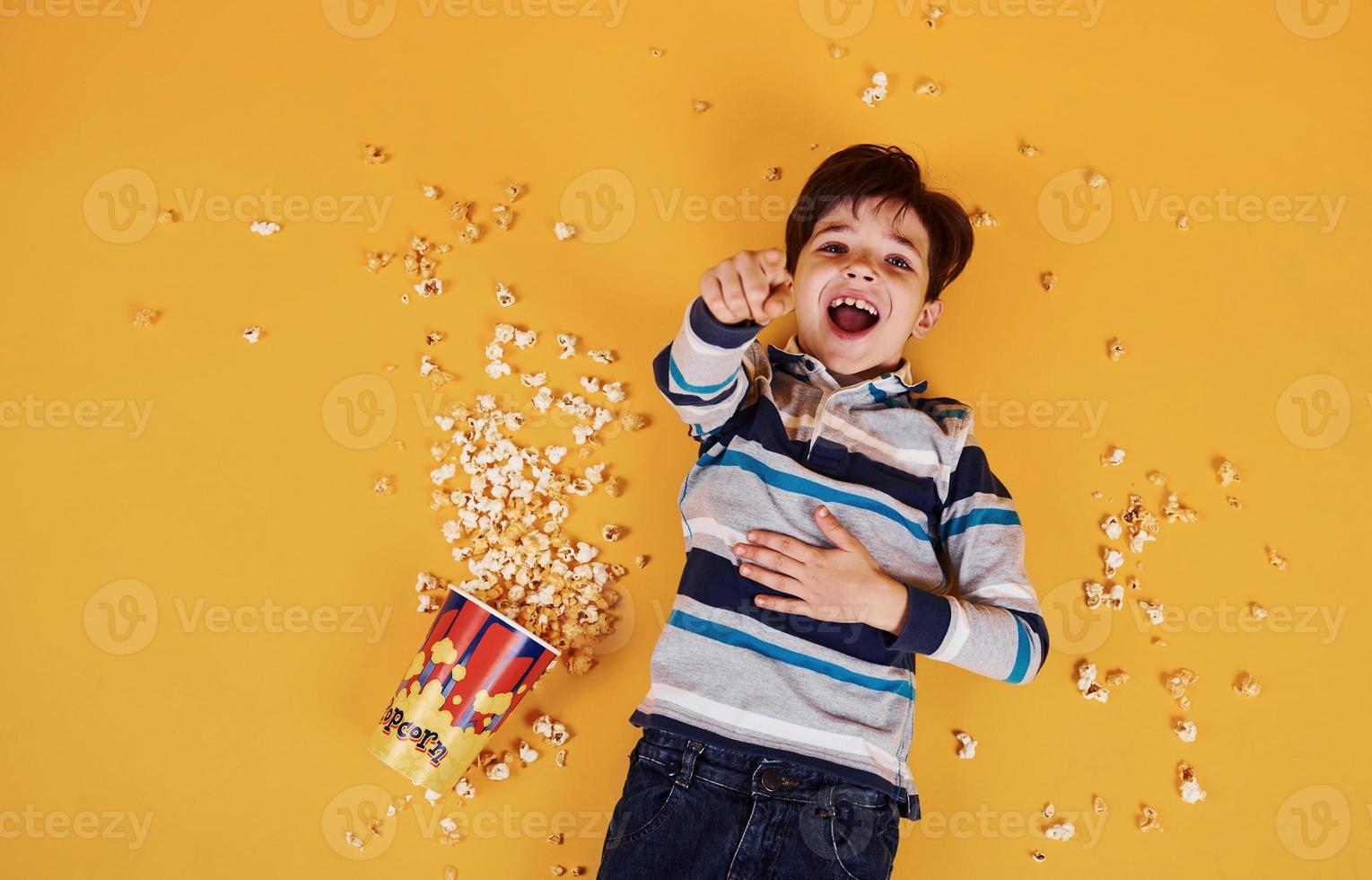 söt liten pojke med popcorn liggande ner på de gul golv foto