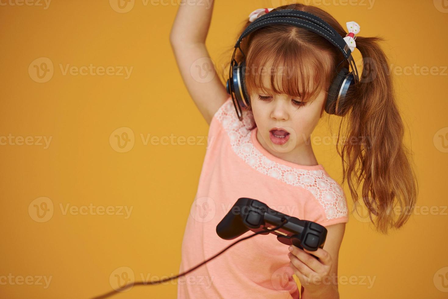 liten kvinna gamer i hörlurar och med joystick i händer spelar video spel mot gul bakgrund foto