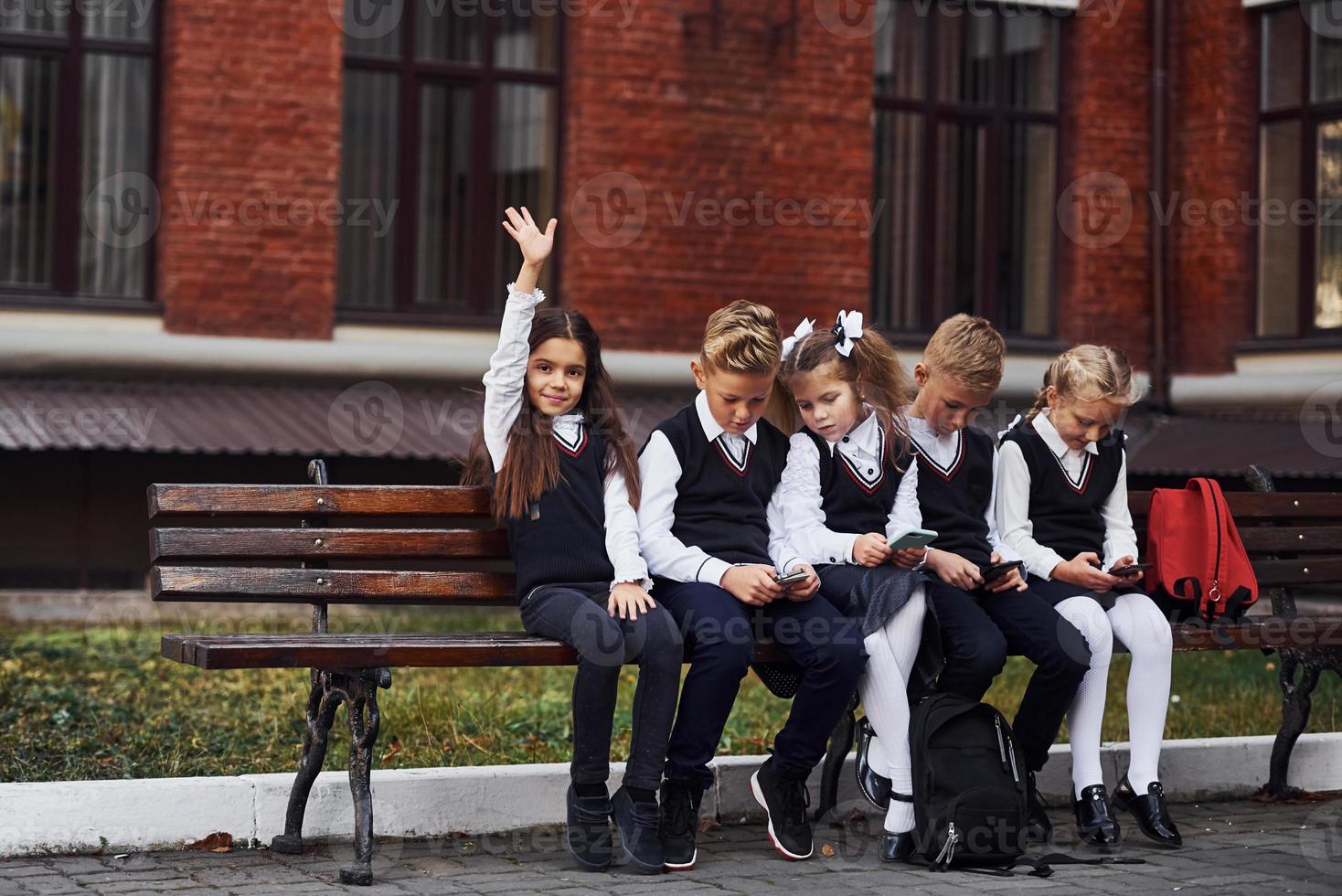 grupp av barn i skola enhetlig sitter på de bänk utomhus tillsammans nära utbildning byggnad foto