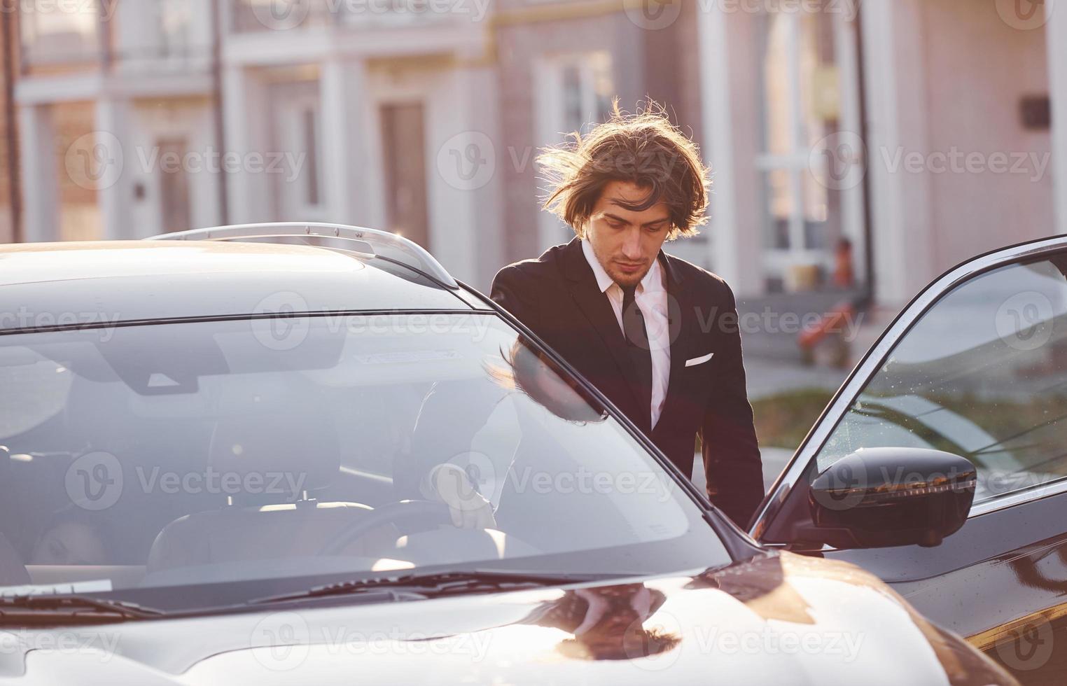 porträtt av stilig ung affärsman i svart kostym och slips utomhus nära modern bil i de stad foto