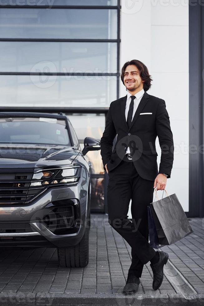 porträtt av stilig ung affärsman i svart kostym och slips utomhus nära modern bil och med handla påsar foto