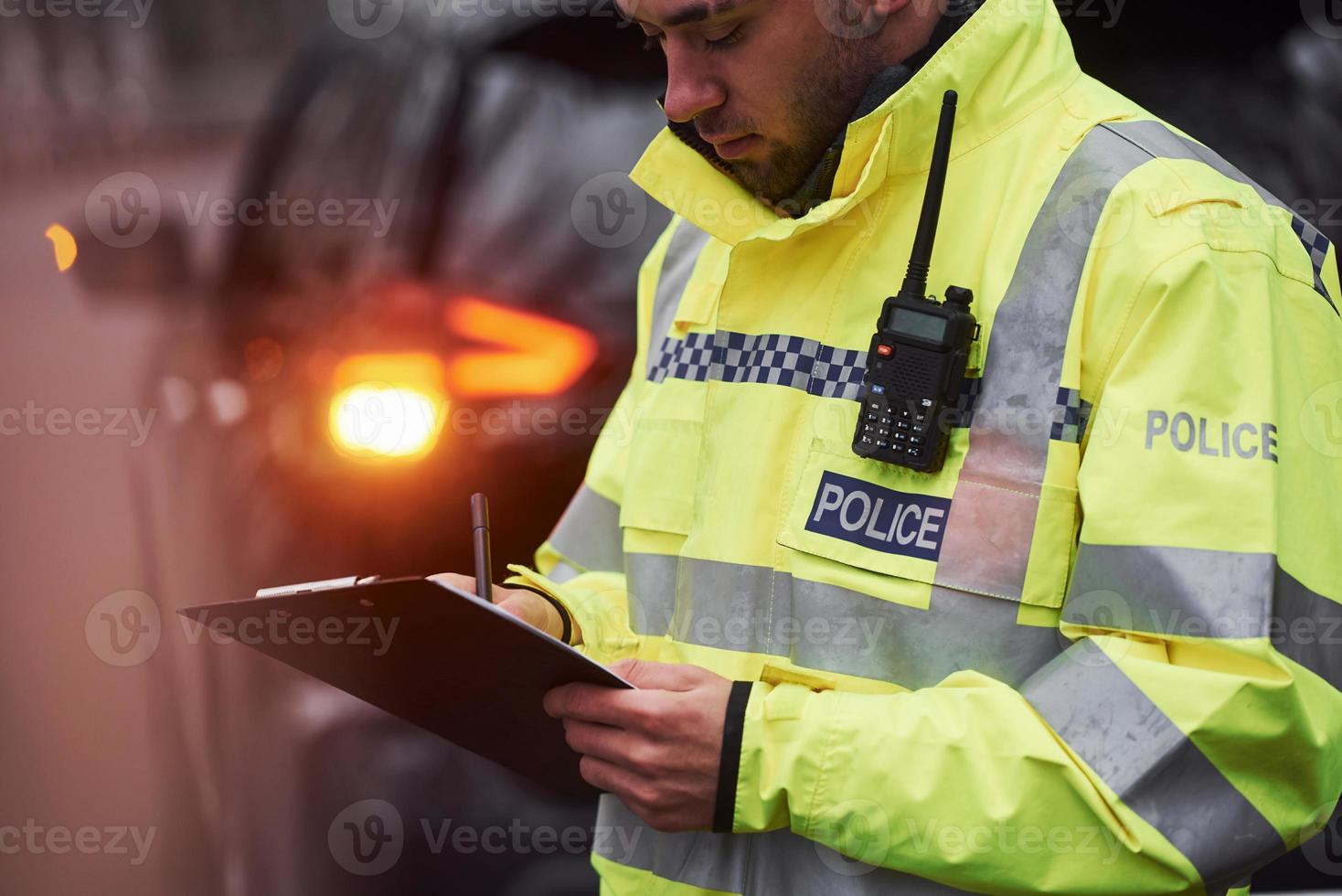 manlig polis officer i grön enhetlig stående med anteckningsblock nära bil foto