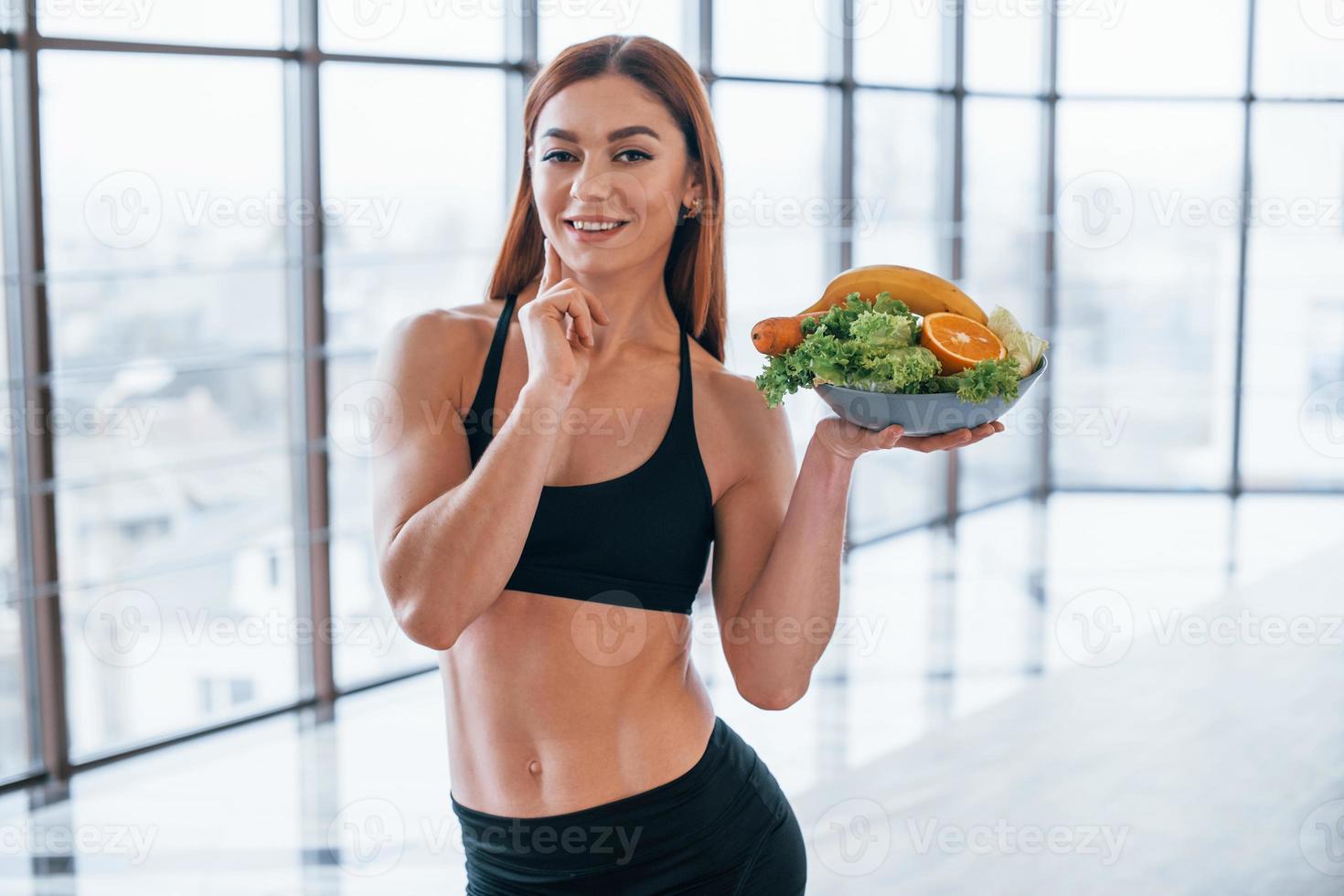 glad sportigt kvinna står inomhus med friska mat i händer foto