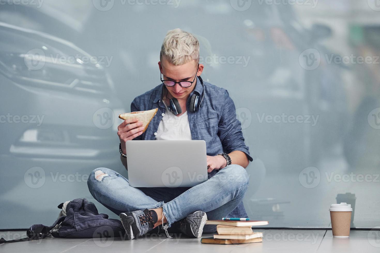 ung hipster kille Sammanträde inomhus nära grå bakgrund med bärbar dator och böcker foto