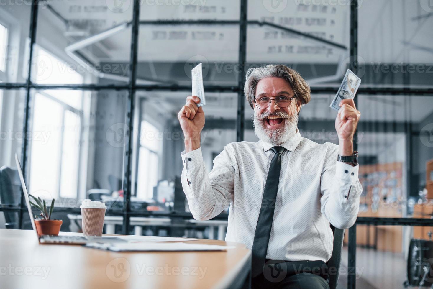 Lycklig mogna affärsman med grå hår och skägg i formell kläder är i de kontor innehav pengar foto