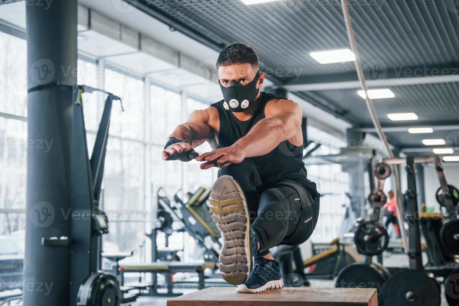 stark ung man i sportigt kläder och skrämmande mask håller på med skjuta på Posten på ett ben i Gym foto