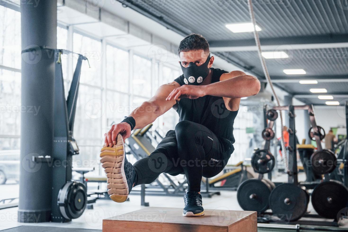 stark ung man i sportigt kläder och skrämmande mask håller på med skjuta på Posten på ett ben i Gym foto