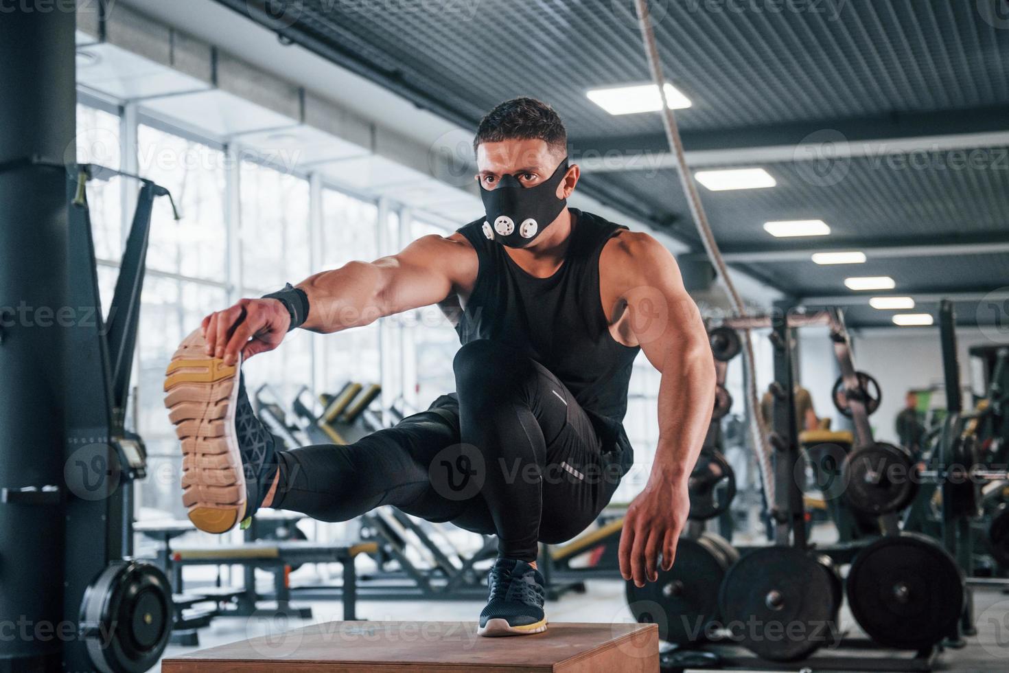 stark ung man i sportigt kläder och skrämmande mask håller på med skjuta på Posten på ett ben i Gym foto