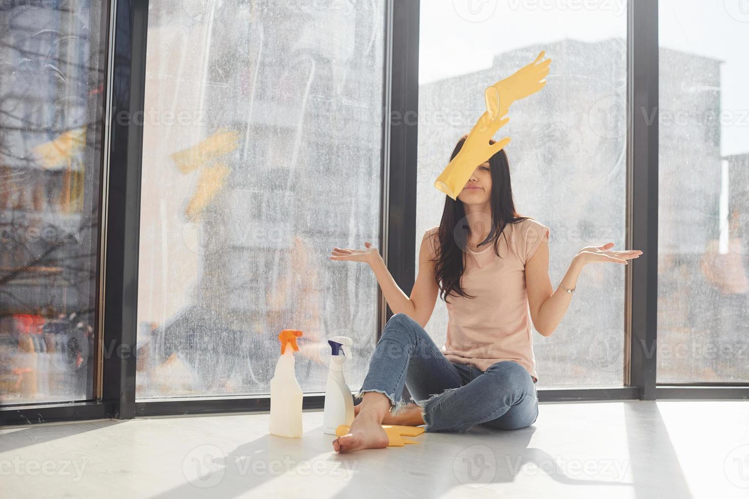 trött kvinna sitter på fönster tröskel med flaskor av rengöring spray och kasta handskar in i de luft foto