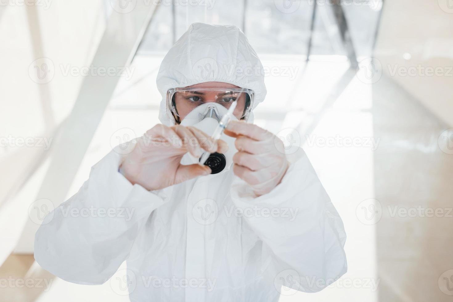 innehar spruta med medicin. kvinna läkare forskare i labb täcka, defensiv glasögon och mask stående inomhus foto
