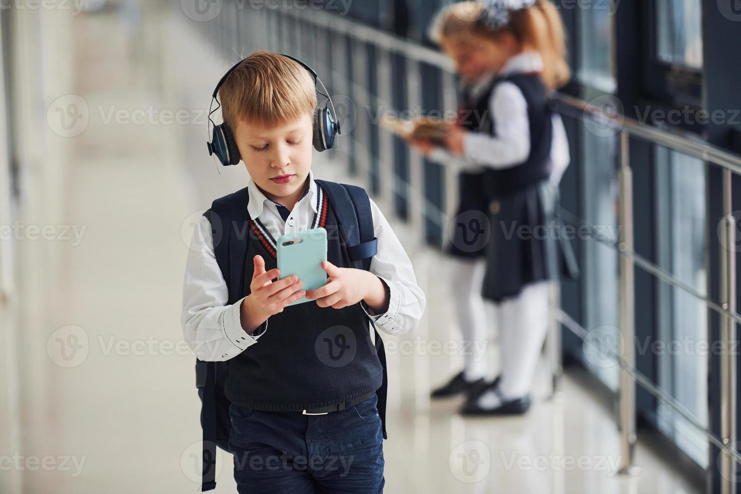 liten pojke med telefon och hörlurar stående om främre av skola barn i enhetlig den där tillsammans i korridor. uppfattning av utbildning foto