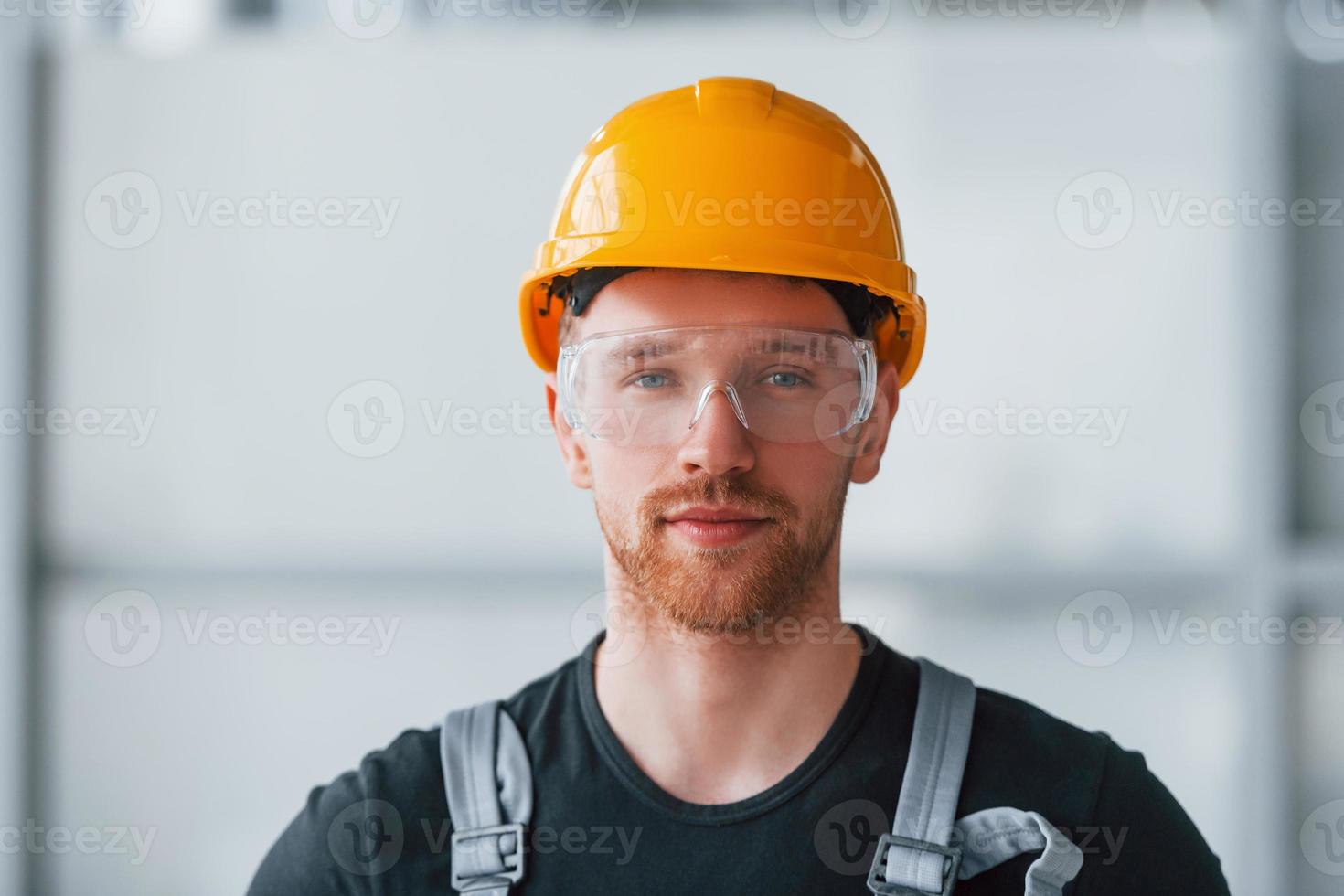 porträtt av man i grå enhetlig, skyddande glasögon och orange hård hatt den där stående inomhus i modern stor kontor på dagtid foto