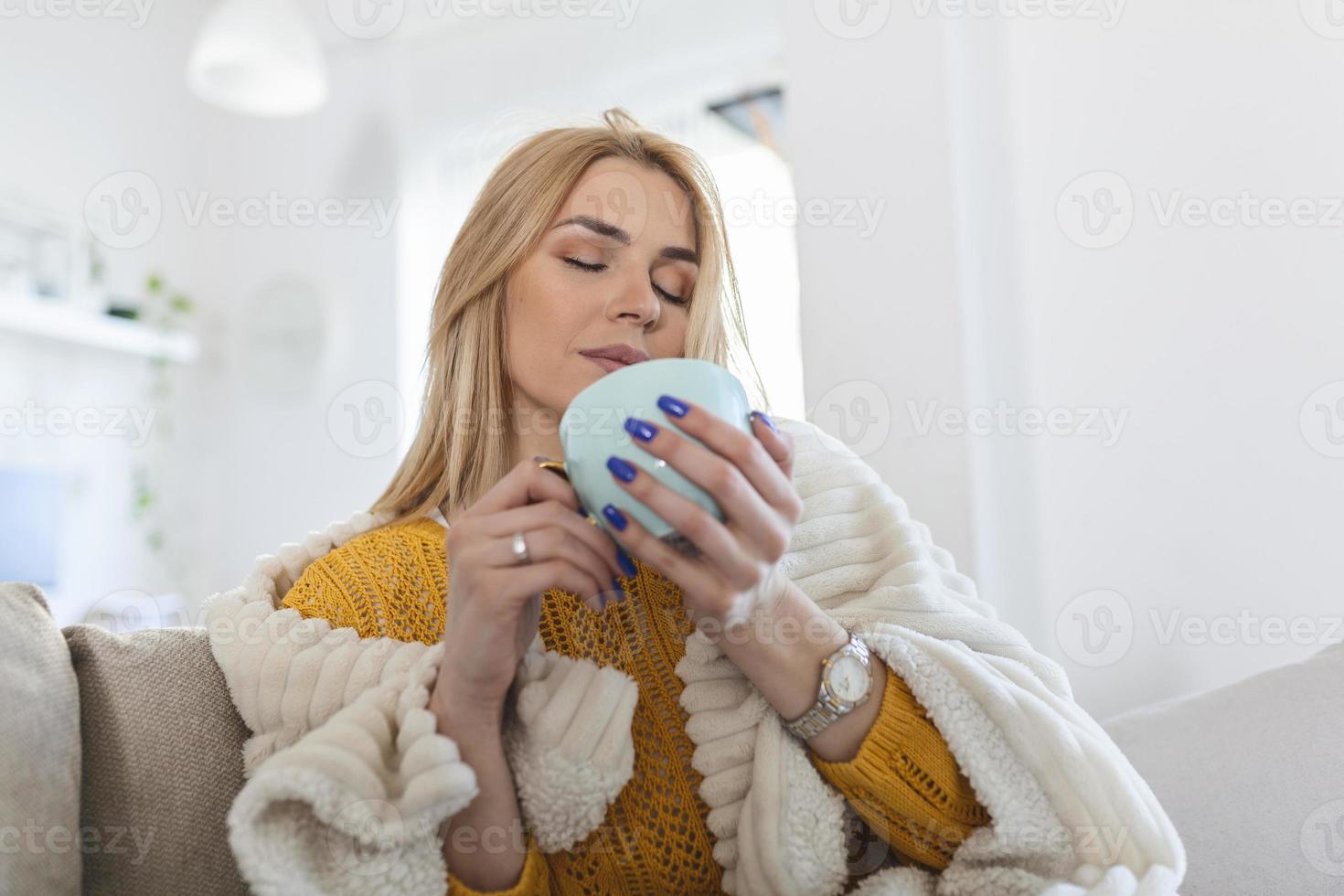 glad ung kvinna dricka kaffe Sammanträde under filt på soffa på hem.ung kvinna är sola under en filt och en kopp av kaffe på en vinter- morgon- på Hem foto