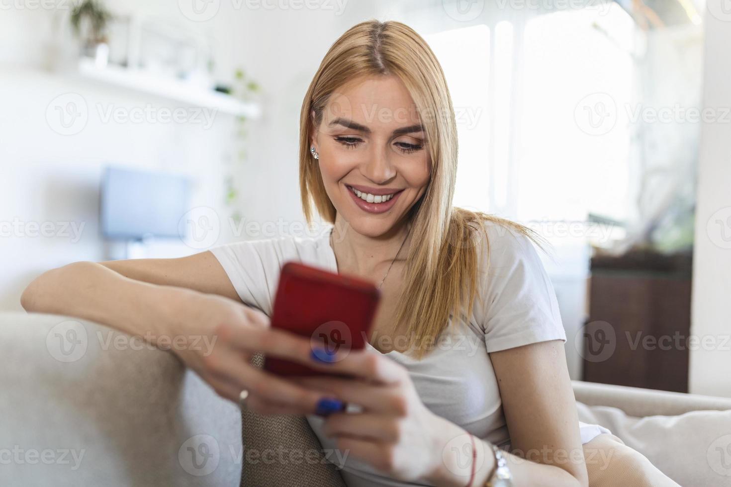 attraktiv leende kvinna använder sig av smart telefon medan Sammanträde på de soffa på Hem. kommunikation och mysighet begrepp. foto