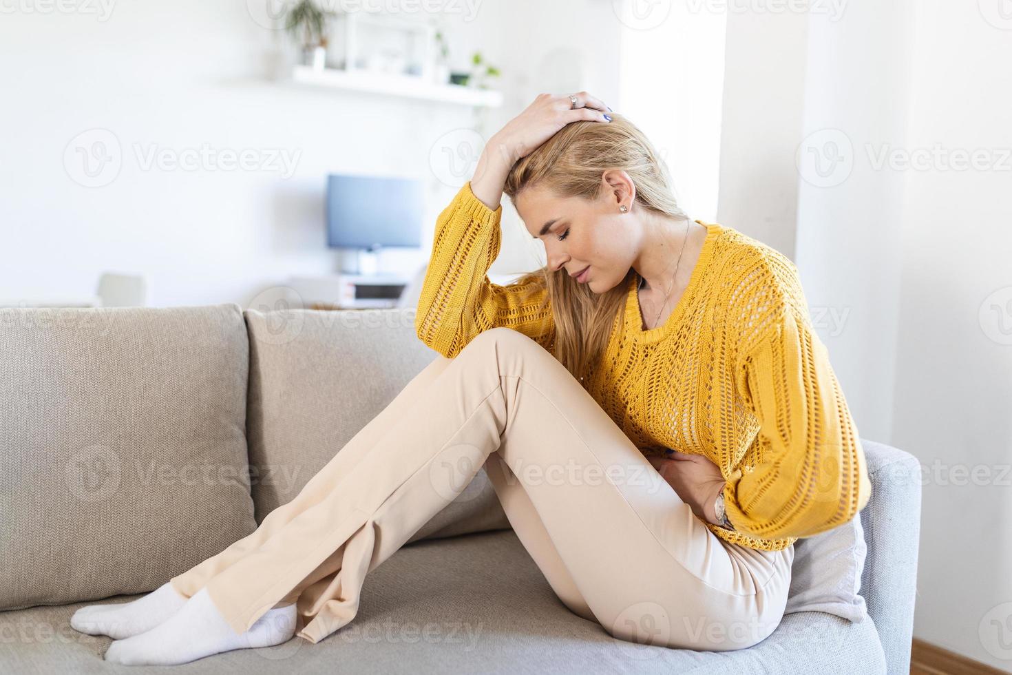 kvinna liggande på soffa ser sjuk i de levande rum. skön ung kvinna liggande på säng och innehav händer på henne mage. kvinna har smärtsam magont på säng, menstruations- period foto