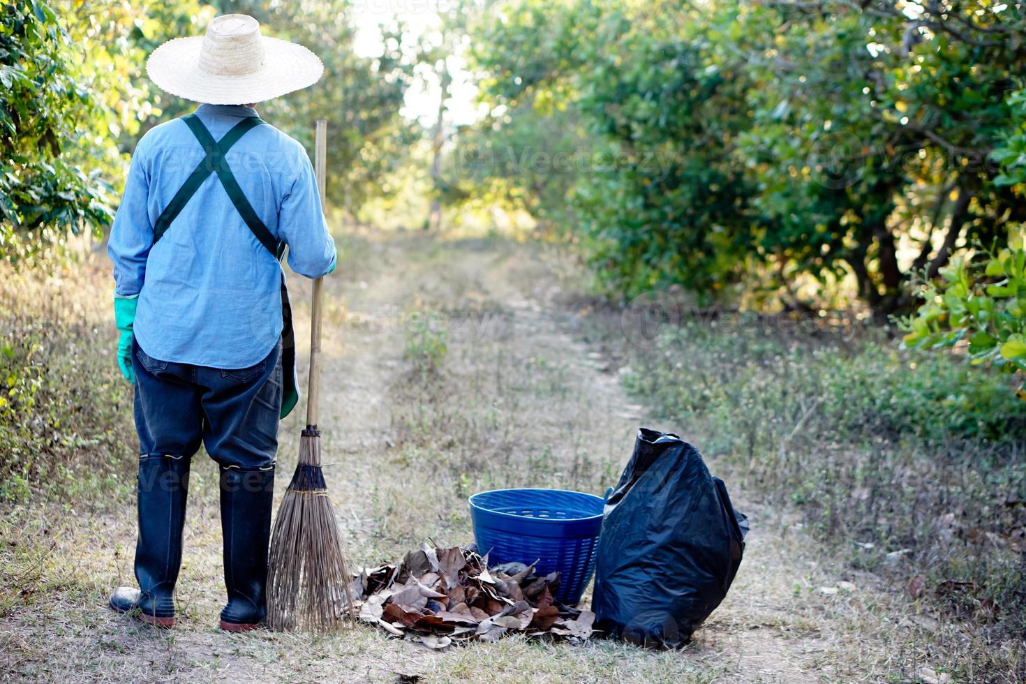 tillbaka se av trädgårdsmästare bär hatt, innehar kvast pinne, står bredvid svart sopor väska och korg i de trädgård. begrepp, sopa torr löv till göra kompost. miljö skydd foto