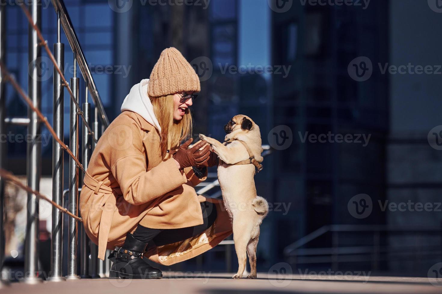 kvinna i värma kläder Sammanträde och ha roligt med henne liten mops hund nära företag byggnad den där på bakgrund foto