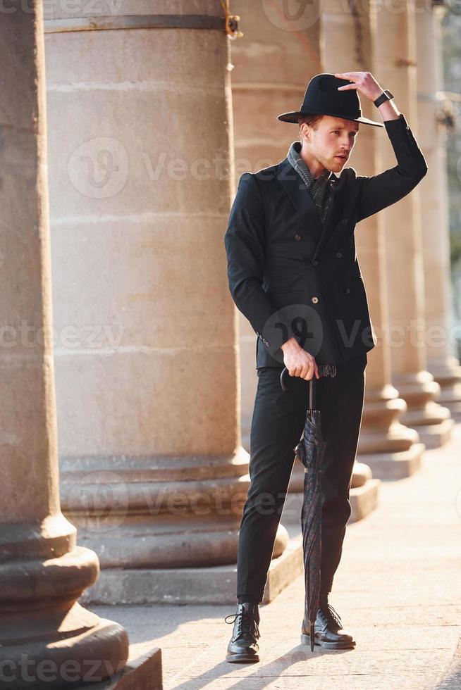 elegant ung man i svart hatt och kläder med paraply i händer utomhus i de stad foto